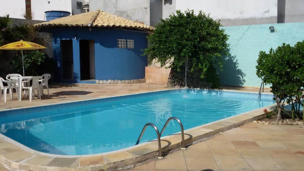 Piscina externa retangular com escada de metal, cercada por um pátio de azulejos, cadeiras, guarda-sol, plantas e um prédio azul, na propriedade no Hotel Pousada Oasis em Conde.