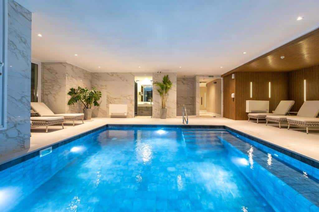 Piscina interna com água azul, cercada por espreguiçadeiras, plantas e paredes de mármore sob luzes brilhantes no teto, no Enjoy Punta del Este, para representar onde ficar em Punta Del Este.