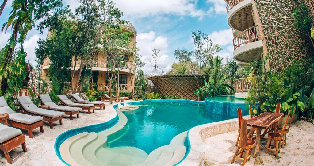 Área da piscina externa com espreguiçadeiras, construções redondas de tecido, árvores e uma mesa de madeira com cadeiras sob um céu nublado, na propriedade do Kan Tulum.