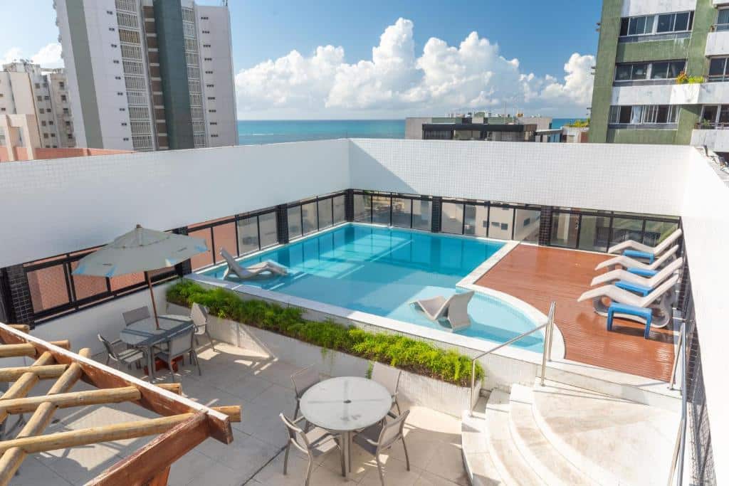 Área da piscina na cobertura com espreguiçadeiras, mesas, guarda-sóis e vista para os prédios da cidade e o oceano ao fundo, na propriedade do Palms Ponta Verde by Tropicalis na praia de Ponta Verde.