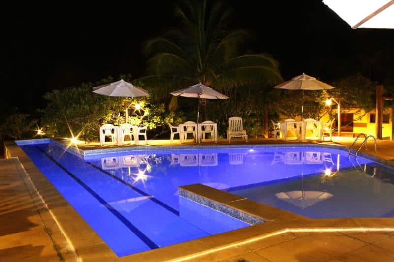 A piscina é iluminada à noite, cercada por espreguiçadeiras e guarda-sóis brancos, com árvores ao fundo, na propriedade da Pousada Bichelenga.