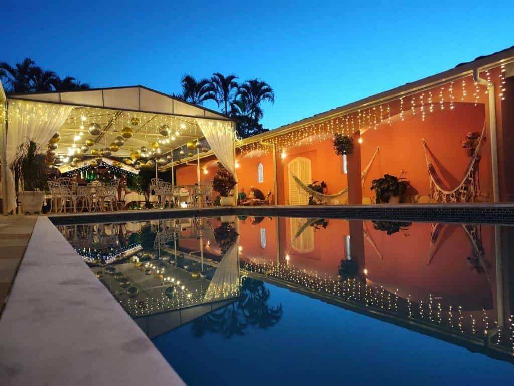 Espaço para eventos ao ar livre com luzes de corda, refletores de piscina, paredes laranja e redes ao entardecer, na propriedade da Pousada Casa Doce Vida.