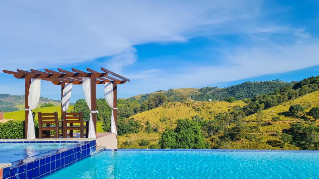 Uma pérgola de madeira com um banco tem vista para uma piscina azul, com colinas verdes e um céu claro ao fundo, na propriedade da Pousada Estância das Montanhas.