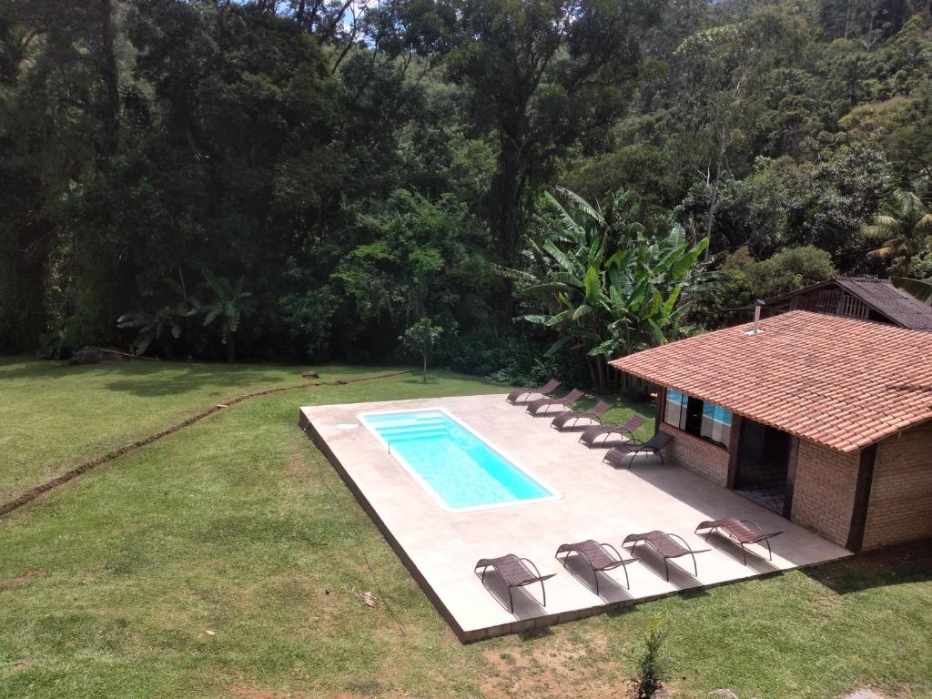 Piscina retangular com espreguiçadeiras ao lado de um prédio de tijolos, cercada por grama e árvores densas, na propriedade da Pousada Pizzaria do Vale.