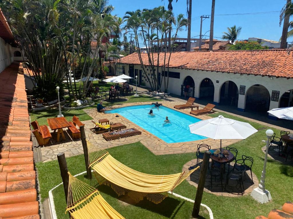 Pátio externo com piscina, espreguiçadeiras, mesas, guarda-sóis, redes e várias pessoas aproveitando a área, na propriedade da Pousada Puerta Del Sol Rio das Ostras.