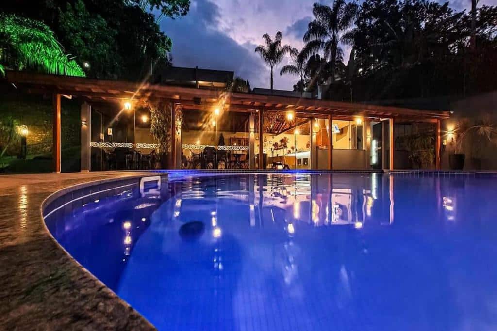 Uma piscina iluminada em frente a um moderno bar ao ar livre e área de lounge ao anoitecer, com árvores ao fundo, na propriedade Pousada Recanto da Cascata.