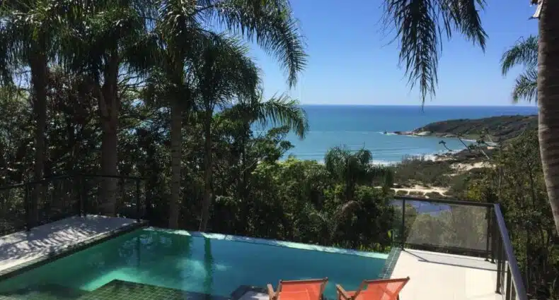 Uma pequena piscina na Pousada The Rosebud com duas espreguiçadeiras laranja tem vista para uma praia tropical e para o oceano, cercada por palmeiras, para representar pousadas na Praia do Rosa.