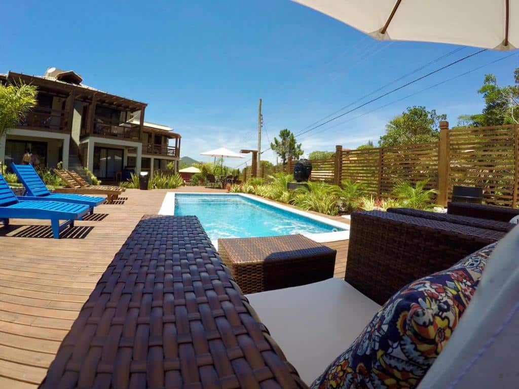 Área da piscina externa com móveis de vime, espreguiçadeiras azuis, deck de madeira e vegetação sob um céu claro, na propriedade da Pousada Vivenda do Rosa.