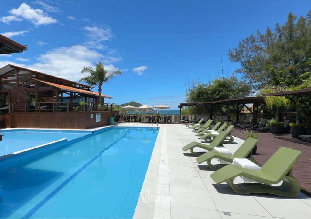 Piscina externa com espreguiçadeiras verdes, deck de madeira e um prédio sob um céu parcialmente nublado, na propriedade do Village Praia do Rosa.
