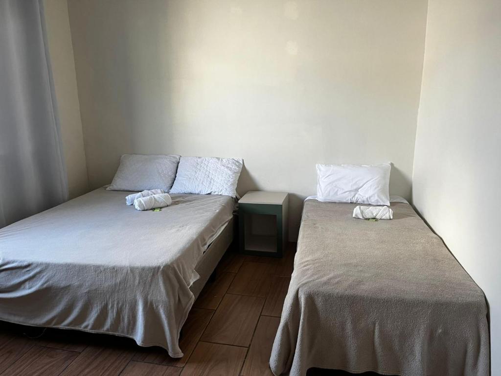 Duas camas bem arrumadas com toalhas brancas em cada uma e uma pequena mesa de cabeceira entre elas em um quarto simples e de cor clara, na POUSADA BLUE GARDEN, para representar pousadas em Lages.