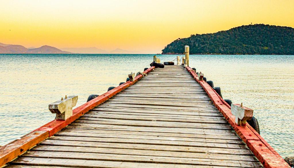 Um píer de madeira se estende sobre águas calmas em direção a uma colina distante coberta de árvores ao pôr do sol, na Pousada Casa do Oscar.