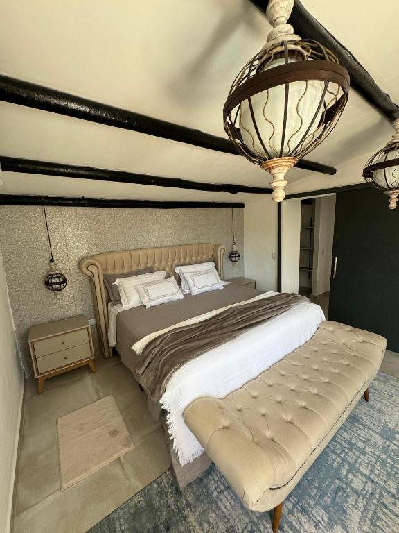 Uma cama bem arrumada com cabeceira bege, duas mesinhas de cabeceira, luzes penduradas e um banco aos pés da cama, em um quarto na Pousada Chalé da Montanha São Tomé das Letras.