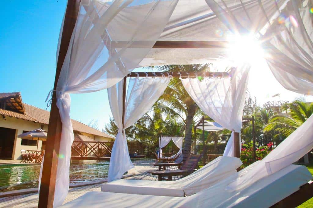 Espreguiçadeiras brancas descansam sob um dossel com cortinas transparentes ao lado de uma piscina, com palmeiras e o sol brilhando ao fundo, na Pousada Coqueiro Beach.