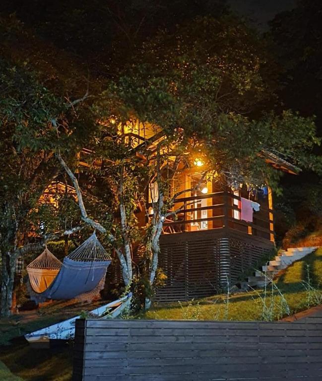 Uma cabana de madeira com iluminação aconchegante à noite, cercada por árvores, com duas redes penduradas nas proximidades e um pátio gramado inclinado, no Farol do Arvoredo Cabanas, para representar pousadas em Governador Celso Ramos.