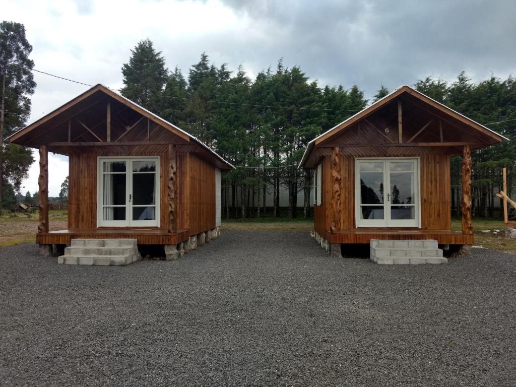 Duas pequenas cabanas de madeira com degraus na frente e janelas grandes, situadas no cascalho com árvores ao fundo sob um céu nublado, na propriedade da Pousada Recanto do Lago em Cambará do Sul.