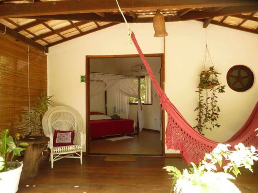 Pátio coberto com cadeira de vime, rede vermelha, vasos de plantas e porta aberta que leva a um quarto com cama de dossel, na propriedade da Pousada Roana.