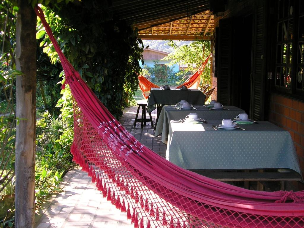 Uma rede vermelha está pendurada em uma varanda perto de mesas com toalhas de mesa verdes e xícaras de chá sob um telhado de telhas, na propriedade da Pousada Rosa & Canela.