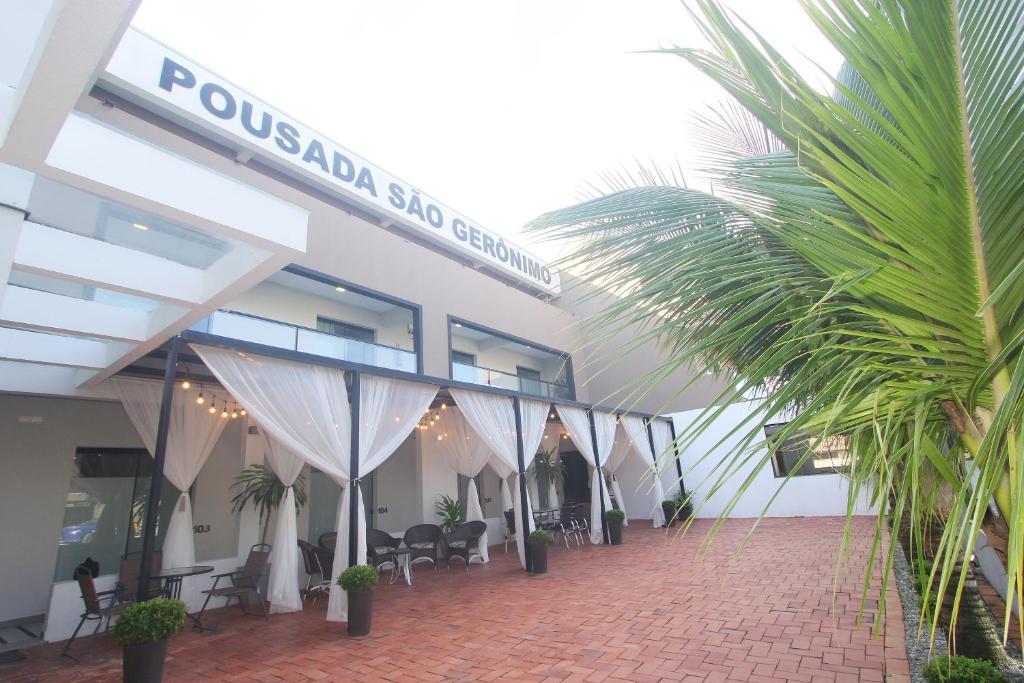 Um pátio de tijolos com vasos de plantas e assentos ao ar livre em frente à Pousada São Gerônimo, um prédio de dois andares com cortinas brancas.