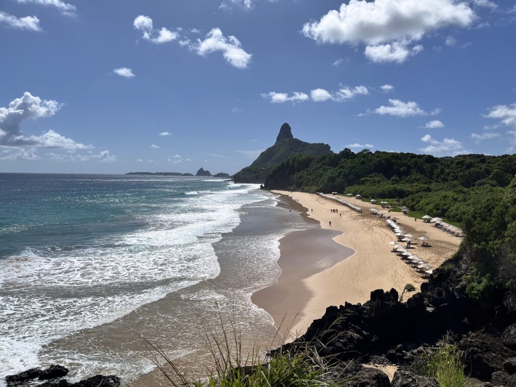 Uma praia de areia serpenteia ao longo da costa com guarda-sóis, colinas verdes e um pico rochoso sob um céu parcialmente nublado.