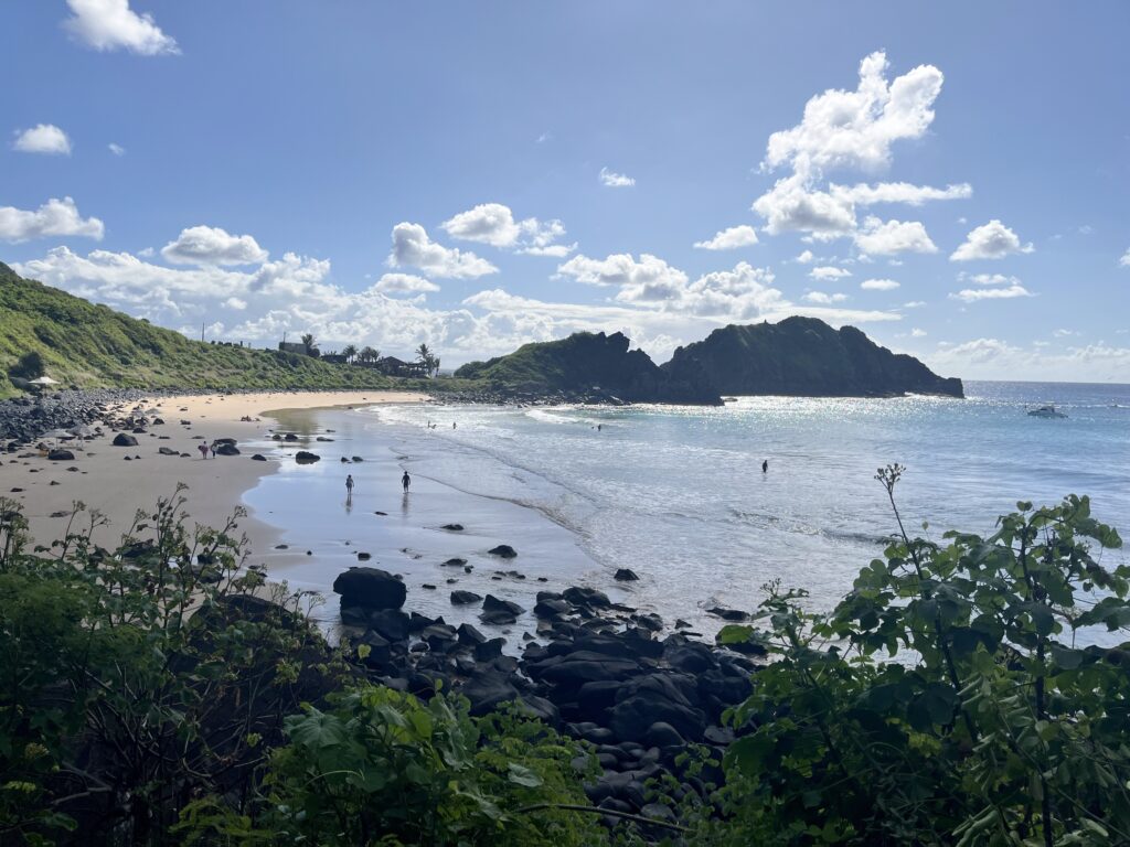Uma praia de areia com pedras espalhadas, colinas verdes e algumas pessoas perto da costa sob um céu parcialmente nublado.