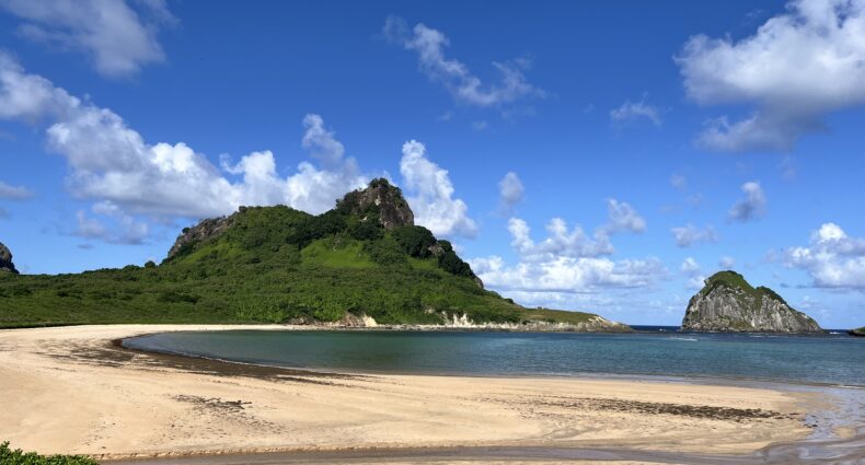 Praia de areia com uma colina verde, águas azuis calmas e uma ilhota rochosa sob um céu claro com nuvens dispersas.