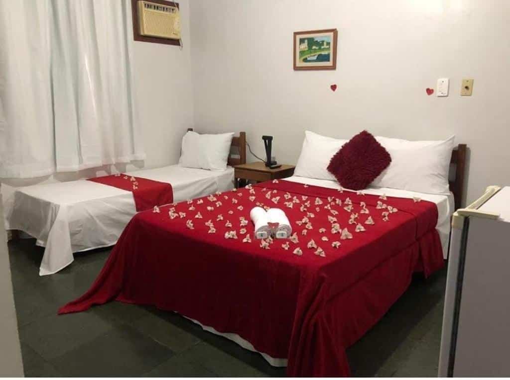 Quarto na Pousada da Jaqueira - EPP, com uma cama de solteiro e uma cama de casal, ambas com mantas vermelhas e pequenas decorações; toalhas na cama de casal, para representar pousadas em Rio das Ostras.