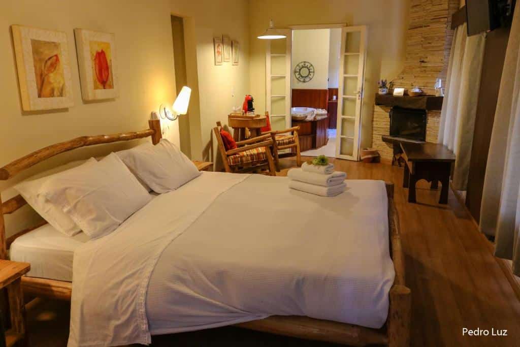 Quarto aconchegante da Pousada Villa Luna, uma das pousadas em Penedo, com uma cama de madeira, roupa de cama branca, uma pequena mesa com cadeiras e uma porta aberta que dá para um banheiro.