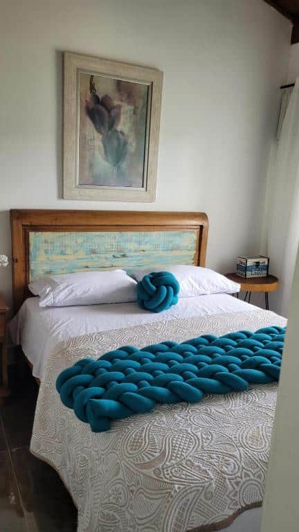 Uma cama bem arrumada no Apto Duplex com Mezanino com roupas de cama brancas, detalhes em tricô de cor azul-petróleo e uma obra de arte emoldurada acima da cabeceira de madeira, para representar airbnb na Praia da Baleia.