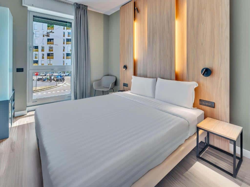 Quarto moderno no B&B Hotel Milano Portello com uma cama de casal, duas mesas de cabeceira, uma cadeira e uma grande janela com vista para a cidade, para representar hotéis em Milão.