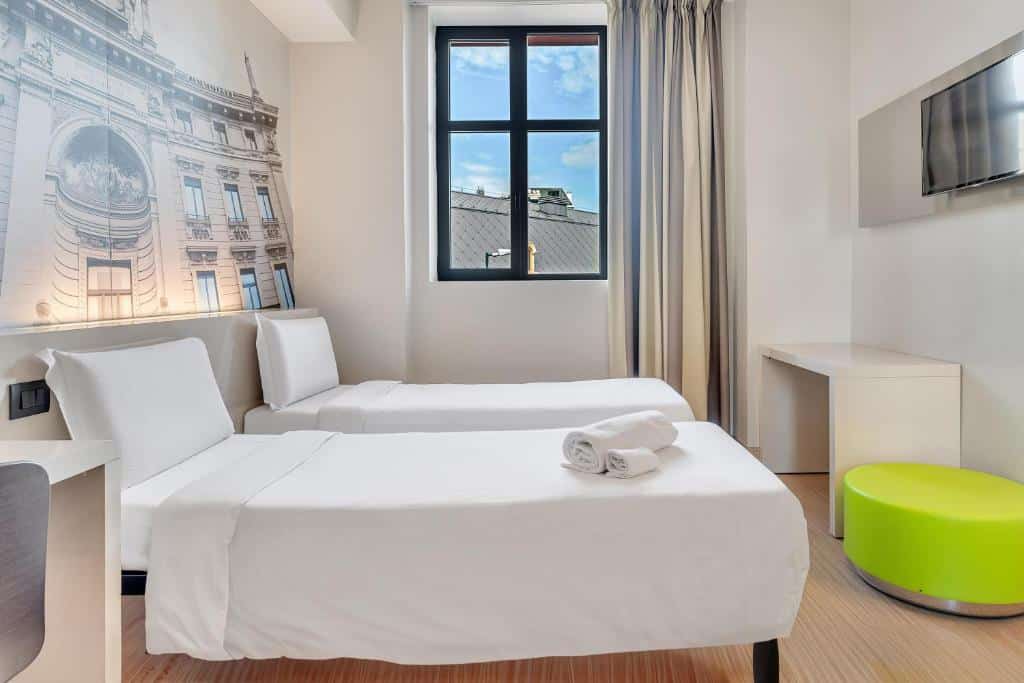Um quarto no B&B Hotel Milano San Siro com duas camas de solteiro, uma janela, uma TV na parede, uma escrivaninha, uma cadeira e um banco verde redondo, para representar hotéis Mercure em Milão.