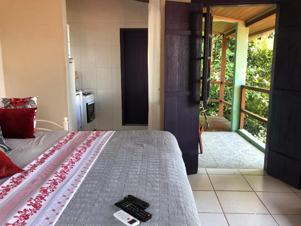 Uma cama com roupa de cama cinza e vermelha, controles remotos em cima, ao lado de uma porta aberta que dá para uma varanda com vegetação do lado de fora, no Bergamota, para representar airbnb em Ilha Grande.