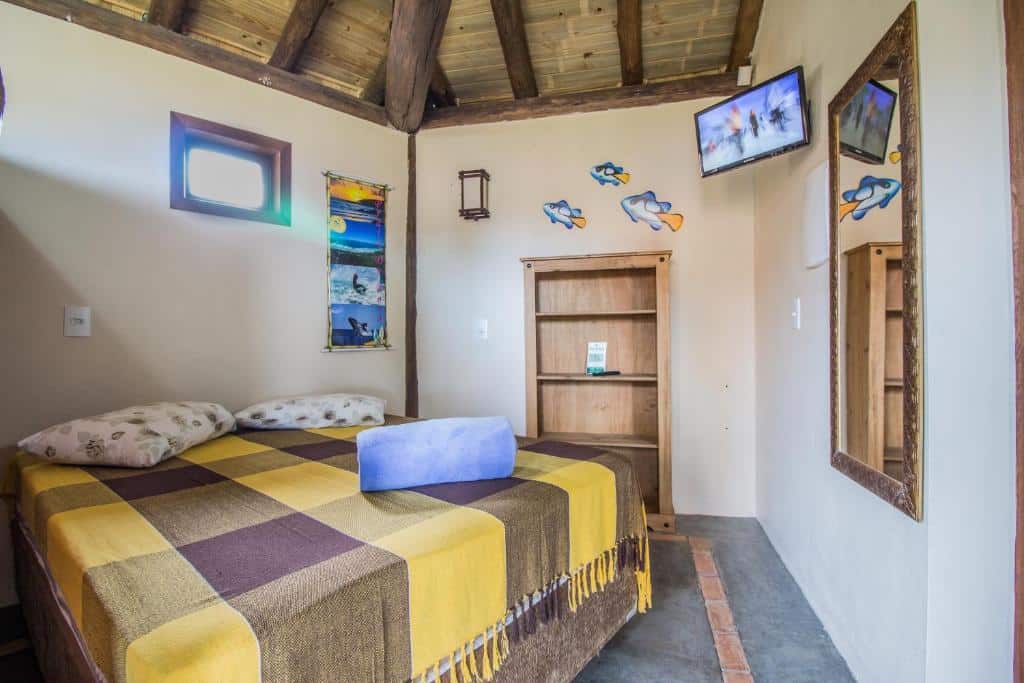 Quarto com uma cama de casal, cobertor amarelo e marrom, arte na parede, TV, espelho, toalha e estante de madeira, na Cabanas do Rosa, para representar pousadas na Praia do Rosa.