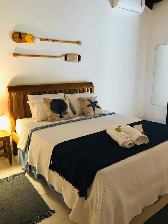 Uma cama bem arrumada com travesseiros com tema de conchas, uma manta azul-marinho e dois remos montados na parede acima da cabeceira, no Canto do Una- hospedagem, para representar pousadas na Barra do Una.