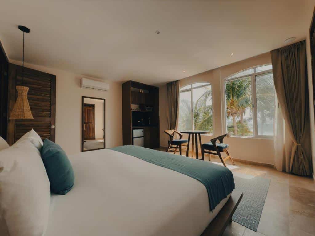 Um quarto na Casa Altamar com uma cama grande, uma mesa pequena com duas cadeiras e janelas grandes com vista para as palmeiras, para representar hotéis em Tulum.