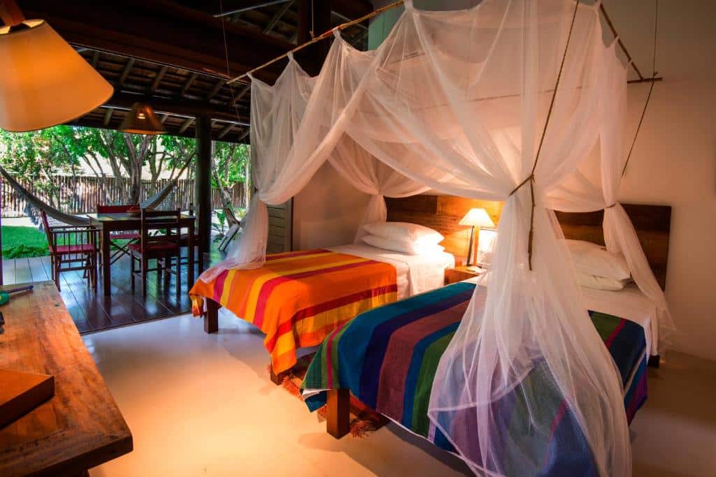 Duas camas com cobertores listrados coloridos e mosquiteiros em um quarto com pátio, rede e iluminação aconchegante, na Casa Sabiá, para representar airbnb em Imbassaí.