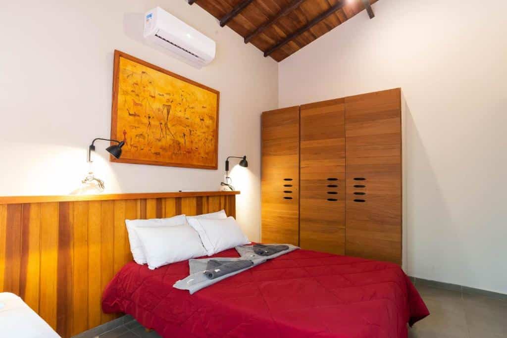 Uma cama com um edredom vermelho, cabeceira de madeira, luminárias de parede, guarda-roupa grande e uma pintura laranja acima da cama, na Casa Tubarão - A Melhor, para representar airbnb na Praia da Baleia.