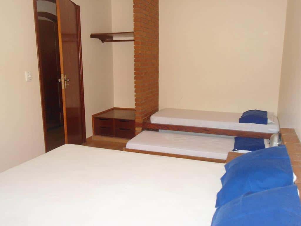 Quarto no Chalés Estrela Do Mar com uma cama de casal e duas de solteiro, com roupa de cama branca e travesseiros azuis, há também um pequeno armário de alvenaria com cabideiro, para representar pousadas na Praia do Lázaro