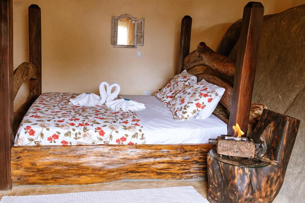 Uma cama de madeira rústica com roupa de cama floral, cisnes de toalha e uma pequena lareira em um suporte de madeira em um quarto aconchegante, nos Chales Sol Paraty, para representar chalés românticos no Rio de Janeiro.