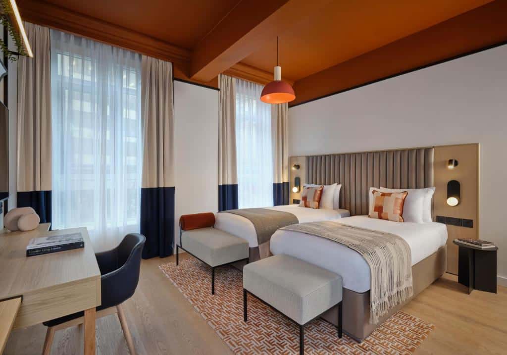 Quarto no Citadines Holborn - Covent Garden London com duas camas de solteiro, uma escrivaninha com cadeira, janelas grandes com cortinas e decoração em tons quentes, para representar airbnb em Londres.