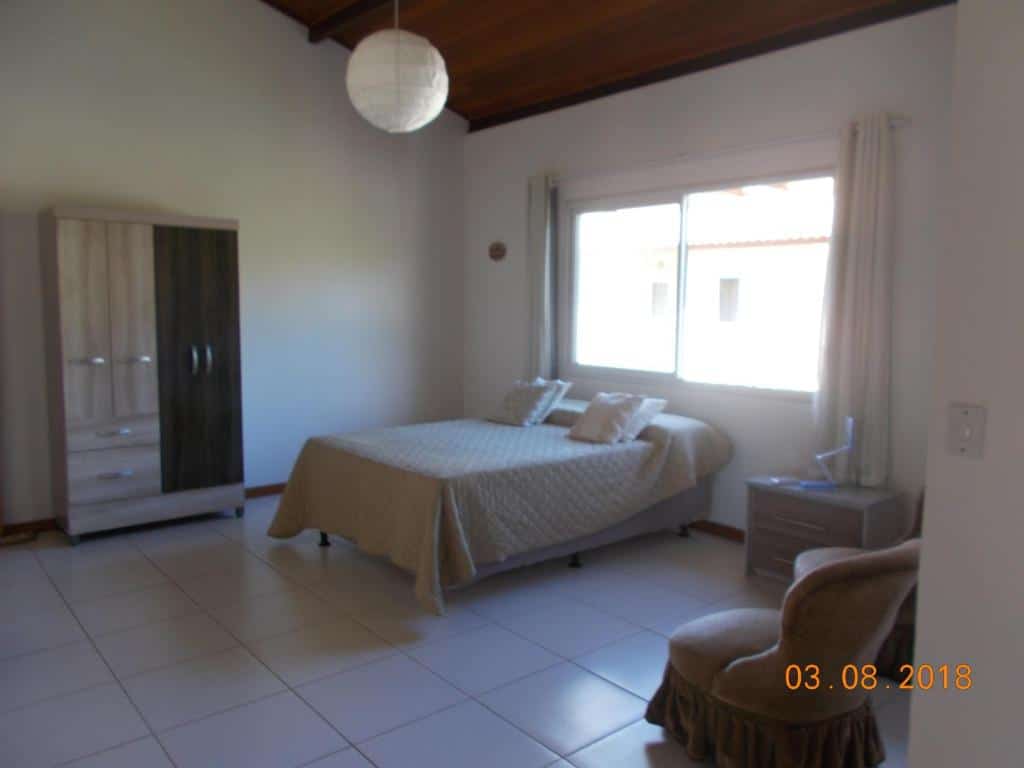 Quarto simples no Cote Dazur Residence com uma cama, guarda-roupa, mesa de cabeceira, cadeira e uma grande janela que permite a entrada de luz natural, para representar pousadas em Imbassaí.