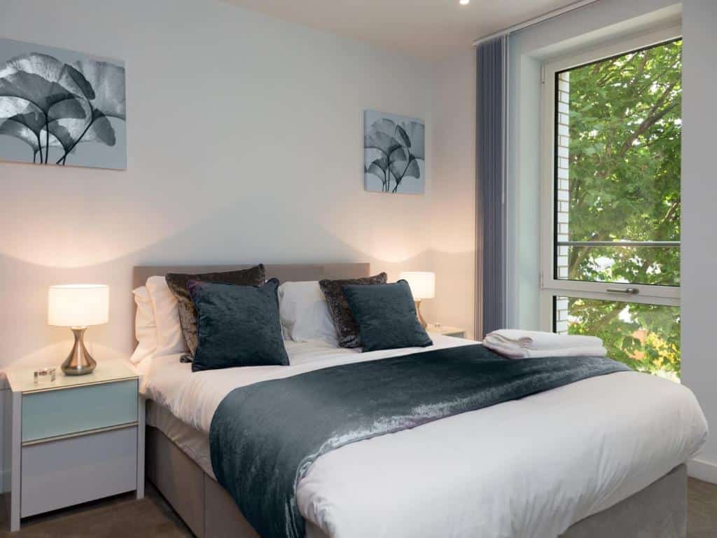 Uma cama bem arrumada com roupa de cama cinza e branca no Deluxe Central London Apartment, duas mesas de cabeceira com abajures e uma janela com vista para as árvores, para representar airbnb em Londres.