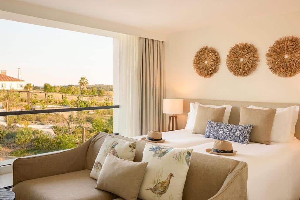 Um quarto no Evora Farm Hotel & Spa com duas camas, decoração bege, um sofá, uma janela grande e arte na parede acima das camas, com vista para um jardim, para representar onde ficar em Évora.