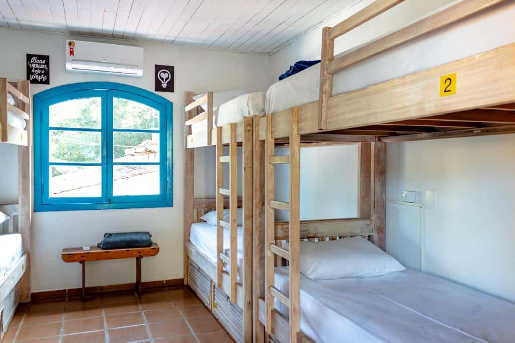 Um pequeno quarto no Flow Hostel Juquehy com dois beliches de madeira, uma janela azul, um banco e paredes brancas com decorações mínimas.