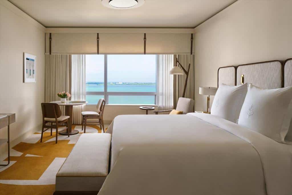 Quarto luxuoso no Four Seasons Hotel Miami com decoração moderna, cama de casal, ampla janela com vista para o mar, uma pequena mesa com duas cadeiras, uma poltrona e um abajur de chão.
