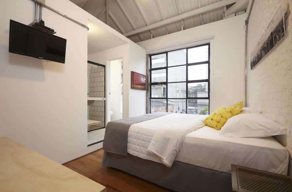 Um pequeno quarto no Guest Urban Hotel Design Pinheiros com uma cama de casal, TV na parede, janela grande e vista para um banheiro com chuveiro de azulejos, para representar hotéis perto do Allianz Parque.