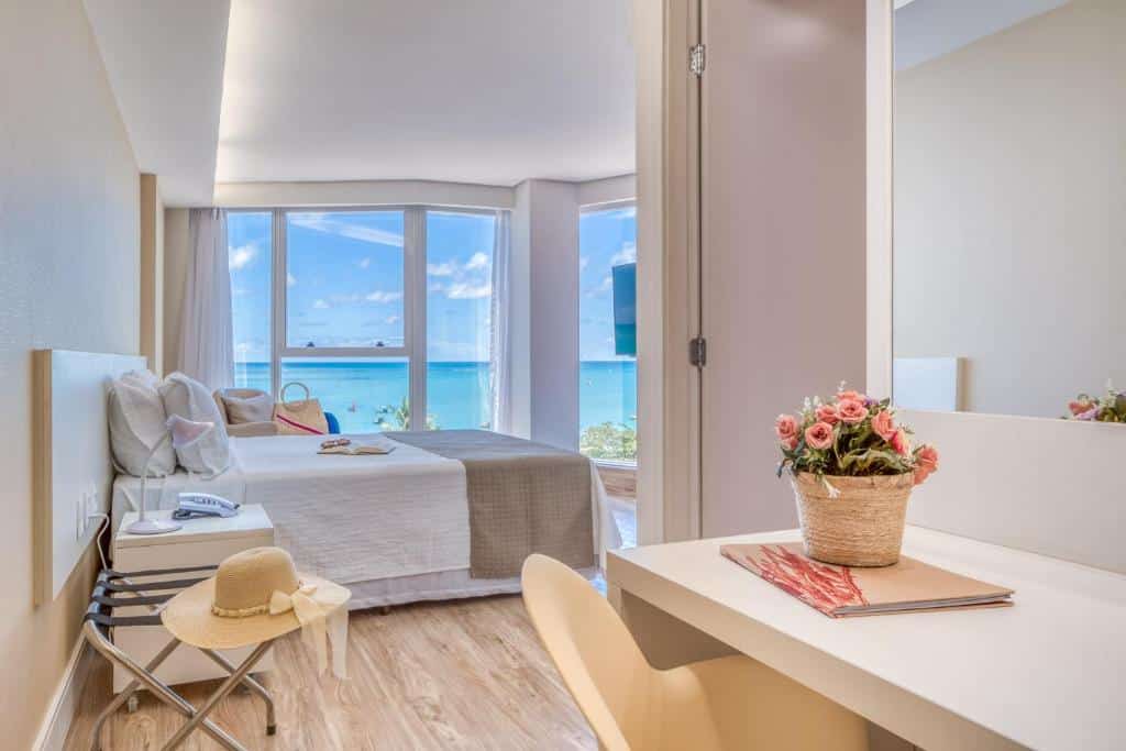 Um quarto no Hotel Brisa Suítes na Praia de Pajuçara, bem iluminado com cama, escrivaninha e grandes janelas com vista para o oceano sob um céu parcialmente nublado, para representar onde ficar em Maceió.