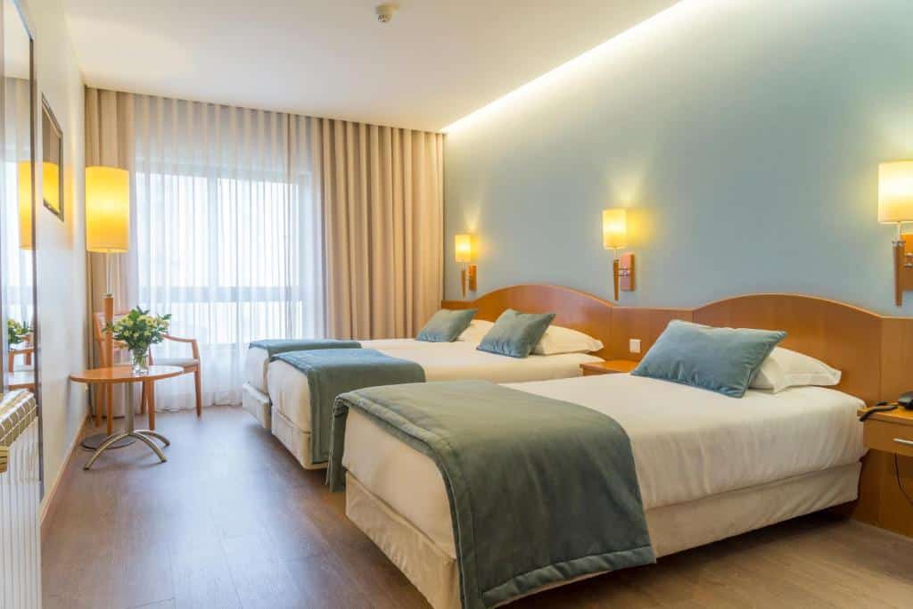 Quarto no Hotel Cinquentenário & Conference Center com duas camas de casal, roupa de cama azul, móveis de madeira, luminárias de parede e uma janela com cortinas transparentes, para representar onde ficar em Fátima.
