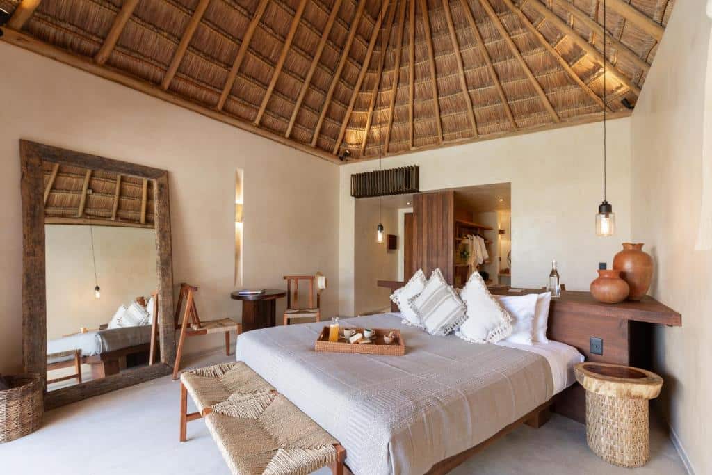 Um quarto rústico no Hotel Ma'xanab Tulum apresenta um telhado de palha, móveis de madeira, um grande espelho e uma decoração em tons neutros com detalhes em cerâmica, para representar hotéis em Tulum.