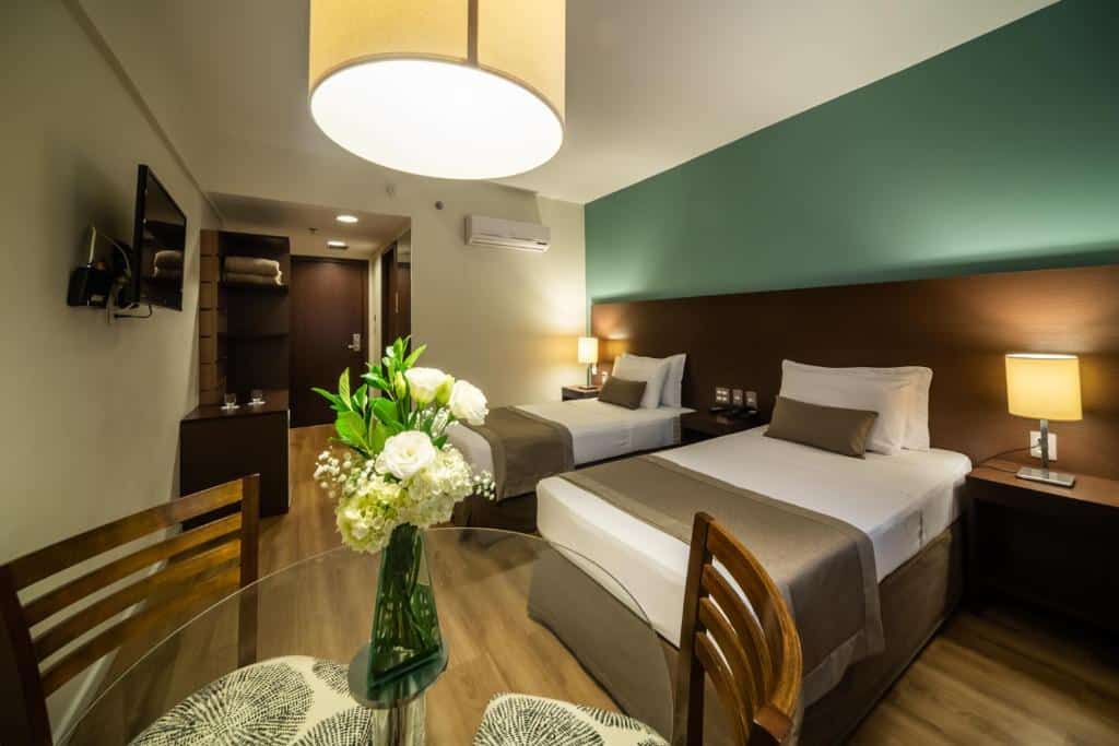 Um quarto no Hotel Panamby São Paulo com duas camas de solteiro, uma mesa de vidro com flores, luminárias e uma TV na parede, para representar hotéis perto do Allianz Parque.