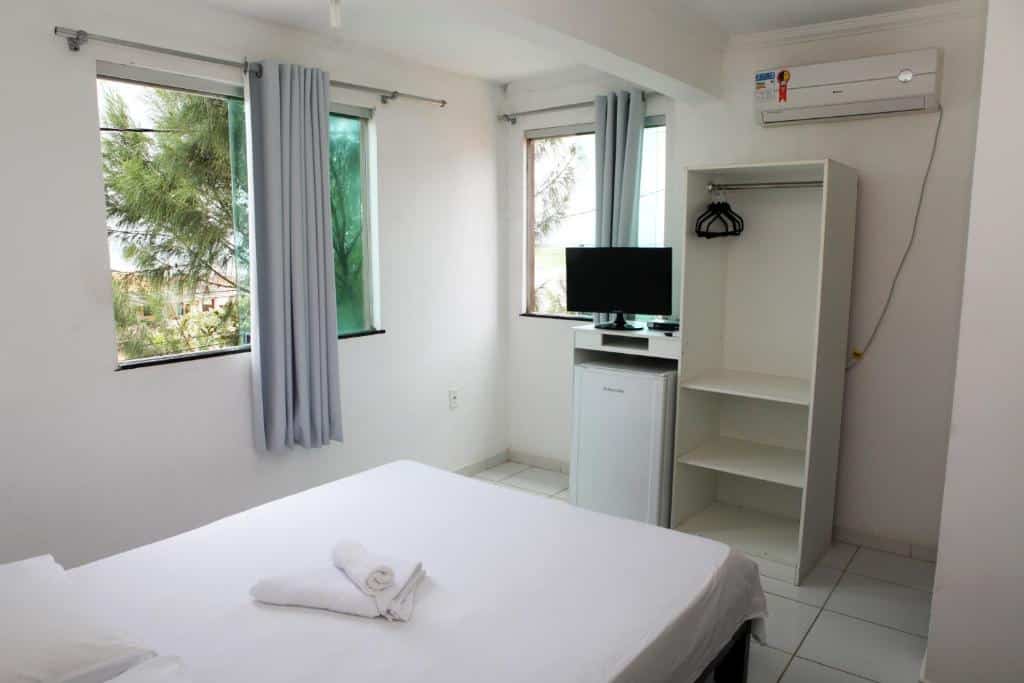 Um quarto simples com uma cama, duas janelas, uma TV, frigobar, armário e ar-condicionado. As toalhas ficam sobre a cama, no Hotel Pousada Oasis em Conde, para representar pousadas na Linha Verde na Bahia.